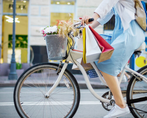 Young woman is cycling around city on white bicycle. Girl in blue dress is going shopping with colorful packages on handlebars of bike. Female retro bicycle with basket with bouquet of flowers.